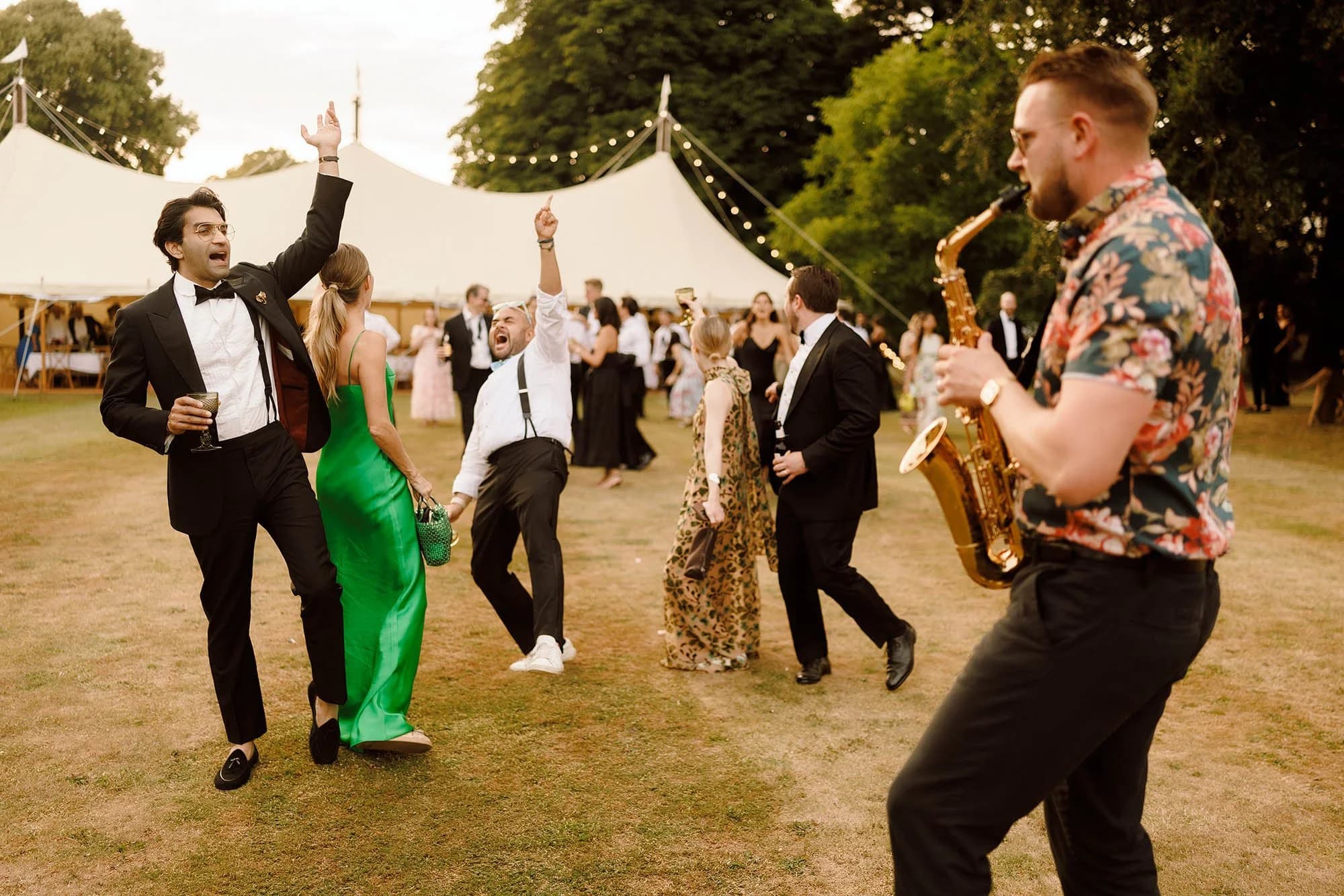 Wedding guests dancing on the grass outside a marquee, a saxophonist playing alongside them